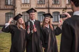 I laureati di successo in abiti accademici sono in possesso di diplomi, guardano la macchina fotografica e sorridono mentre sono in piedi all'aperto, il ragazzo li sta fotografando