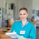 Portrait of young nurse in uniform making notes in medical card and smiling at camera at hospital