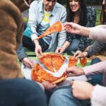 A group of friends sit on the grass and enjoy a large pizza together. The focus of the shot is a close-up of the pizza slices being held by the individuals